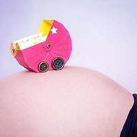 Ein rosafarbener Miniatur-Kinderwagen mit Sternenmotiven steht auf dem runden Babybauch einer schwangeren Frau. Ein kreatives und symbolisches Porträt der Vorfreude auf das Baby, professionell festgehalten von Fotograf Michael Schalansky.